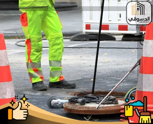 شركة تسليك مجاري في ابوظبي 0545473541 الدسوقي عامل تنظيف المجاري في أبوظبي، مرتديًا زي الأمان، بالقرب من فتحة الصرف الصحي مع الأدوات اللازمة.